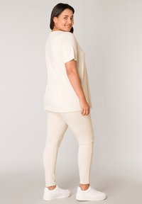 Beige oversized t-shirt met korte mouwen, gecombineerd met luchtige beige leggings en witte sneakers, tegen een neutrale achtergrond.
