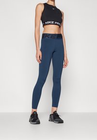 Černý crop top Nike Pro s bílým logem; modré legíny s černým pasem; modelka má na sobě černé sportovní boty. Hladká, přiléhavá látka.
