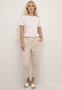 Pantalons beiges ajustés avec plis et taille élastique, associés à un t-shirt en coton blanc uni et des sandales blanches à glisser.