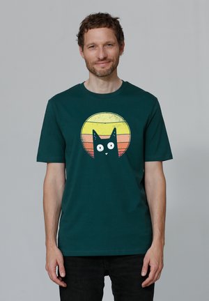 Groene katoenen T-shirt met een grafisch ontwerp van een zwarte kat tegen een ronde zonsondergang in gele en oranje tinten. Korte mouwen, ronde hals.