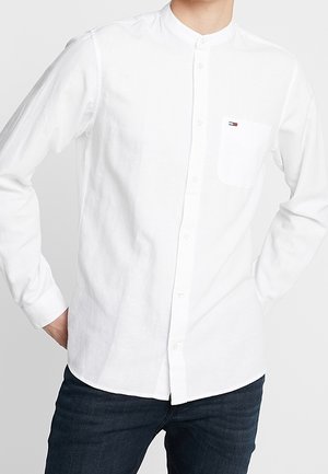 Homme portant une chemise blanche à manches longues avec col mandarin, patte de boutonnage devant et poche poitrine, associée à un jean foncé.