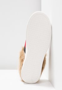 Semelle blanche pour sneaker avec motif triangulaire, vue partielle d’une tige rouge et noire, et bordure en fausse fourrure beige.