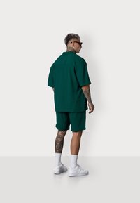 Ein Mann mit einer kurzen, silbernen Frisur trägt ein passendes grünes Outfit und weiße Turnschuhe und steht vor einem schlichten weißen Hintergrund.