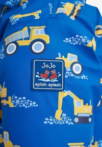 Jachetă impermeabilă albastră cu modele de vehicule de construcție galbene. Accentuata cu un patch branduit care citește "JoJo splish splash."