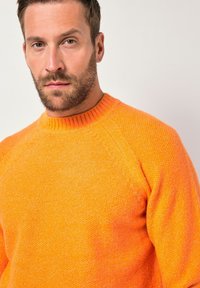 Mies, jolla on ruskeat hiukset ja parta, pukeutunut oranssiin kuvioituun neulepuseroon, katsoo suoraan eteenpäin yksinkertaisen vaalean taustan edessä.