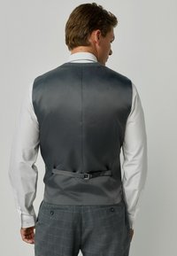 Kulgrå vest med en glat satinfinish, der har en spændejustering bagpå og bæres over en hvid skjorte. Klassisk design.