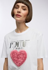 Maglietta bianca in cotone con un design a forma di cuore rosa testurizzato e un testo metallico, "I SOGNI DIVENTANO REALTÀ I’AMOUR amore," sul davanti.