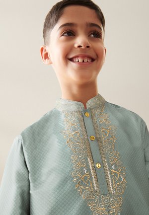 Smiling boy wearing a light blue traditional shirt with gold embroidery and yellow buttons, looking slightly upward against a plain background.