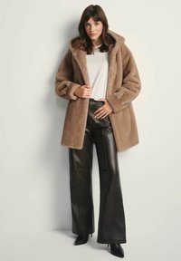 Kobieta ubrana w brązowy płaszcz z sztucznym futrem z kapturem, biały top, czarne skórzane spodnie rozszerzane oraz czarne botki na obcasie z ostrym noskiem, stojąca przy ścianie.