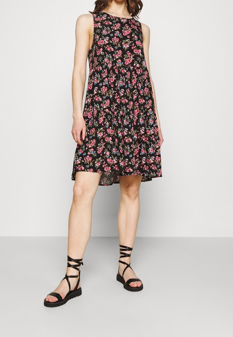 Femme portant une robe noire sans manches avec des motifs floraux roses et bleus et des sandales plates noires à lacets, debout devant un fond uni.