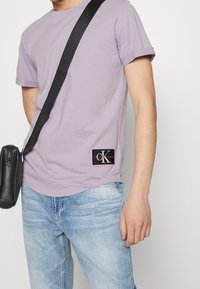 Levandulové bavlněné tričko s krátkým rukávem a ohrnutými manžety, na dolním levém rohu opatřené černou nášivkou s logem Calvin Klein Jeans, v kombinaci s světlými džínami.