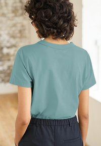 Camiseta de manga corta en color teal con cuello redondo y un tejido suave. Llevada con pantalones negros de cintura elástica, que muestran un ajuste relajado.