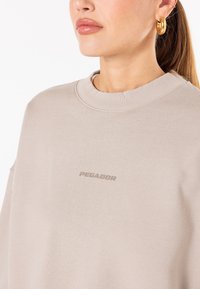 Beige oversized hettegenser laget av myk stoff, med rund hals og subtil "PEGADOR" logo i matchende farge foran.