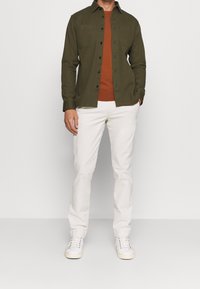 Olivgrünes Hemd mit Knöpfen über einem braunen Pullover, kombiniert mit weißen Hosen und weißen Sneakers. Verfügt über zwei Brusttaschen und schwarze Knöpfe.