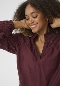 Blouse bordeaux en tissu texturé, avec des détails en dentelle sur les manches, un col froncé et une coupe décontractée.