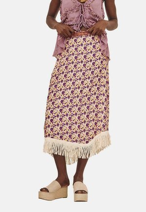 Falda maxi con un patrón floral en púrpura y amarillo, adornada con un fleco crema en el dobladillo. Combinada con una blusa holgada y sandalias de plataforma.