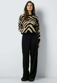 Sieviete, ģērbusies smilšu un melnā zebra svītrainajā džemperī ar melnām platām biksēm, stāv uz vienkārša gaiši pelēka fona.