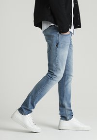 Světlé modré džíny ze denimu se slim střihem, zašedlým předním dílem a kontrastním šitím. Kombinovány s bílými teniskami a černou bundou přes bílé tričko.