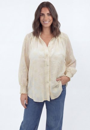 Vrouw met schouderlang bruin haar die lacht, gekleed in een geborduurd roomkleurig blouse en blauwe spijkerbroek, staand met één hand in haar zak.