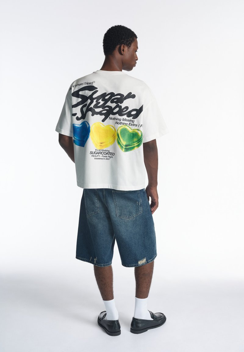 Homme portant un t-shirt blanc oversize avec des motifs colorés de cœurs et du texte, un short en jean, des chaussettes blanches et des chaussures noires, debout de dos.