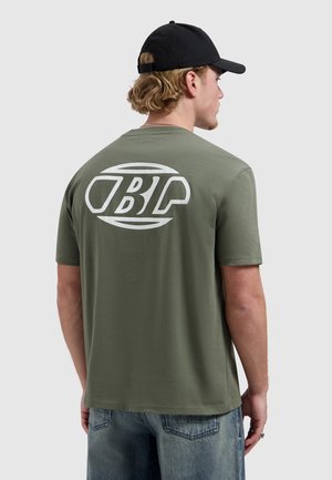 Man draagt een olijfgroen T-shirt met witte gestileerde "BLP"-logo op de achterkant, zwarte pet en blauwe jeans, kijkt van achteren tegen een effen achtergrond.
