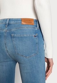 Primer plano de una persona que lleva unos jeans azul claro de Tommy Hilfiger con una etiqueta de cuero marrón de la marca y una camiseta de manga larga blanca con canalé.