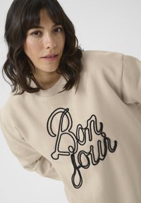Beige sweatshirt featuring a black embroidered "Bonjour" design. The material appears soft with a relaxed fit and ribbed neckline.