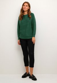 Roheline kampsun pehmest kangast, millel on mugav lõige, ümmargune kaelus ja pikad varrukad, koos mustade kitsaste pükste ja mustade loaferitega.