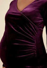 Rochie din catifea violet, având un decolteu adânc în V, design înfășurat și detalii plisate pe lateral, care creează un aspect texturat.