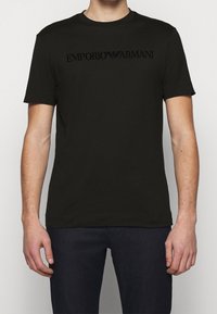 Uomo che indossa una maglietta nera a maniche corte di Emporio Armani e pantaloni scuri, in piedi con le braccia rilassate contro uno sfondo bianco semplice.