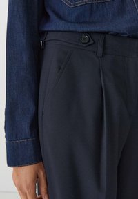 Närbild av svarta skräddarsydda byxor med sidoficka och knappslå, tillsammans med en mörkblå denimskjorta med lång ärm.