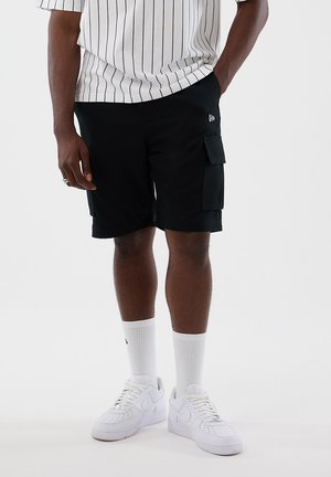 Uomo che indossa pantaloncini cargo neri, calzini bianchi fino al polpaccio, scarpe da ginnastica bianche e una camicia bianca a righe sottili, in piedi con una mano nella tasca.