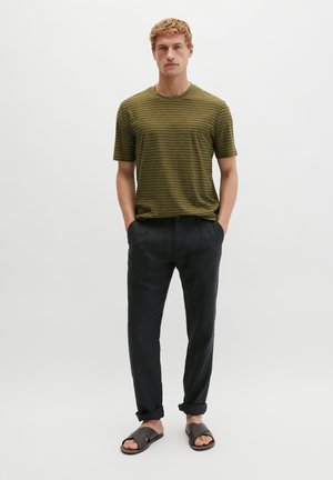 Man standing with hands in pockets, wearing olive green striped t-shirt, black rolled-up pants, and dark sandals against a plain white background.