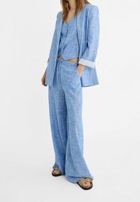 Ljusa blå linnakostym med en figurnära väst med knappar och vida byxor, båda med en texturerad yta och kontrastfärgad midjedetalj.