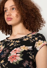 Blusa preta floral com decote redondo, mangas curtas e padrões intrincados de flores em rosa, amarelo e verde sobre um tecido texturizado.