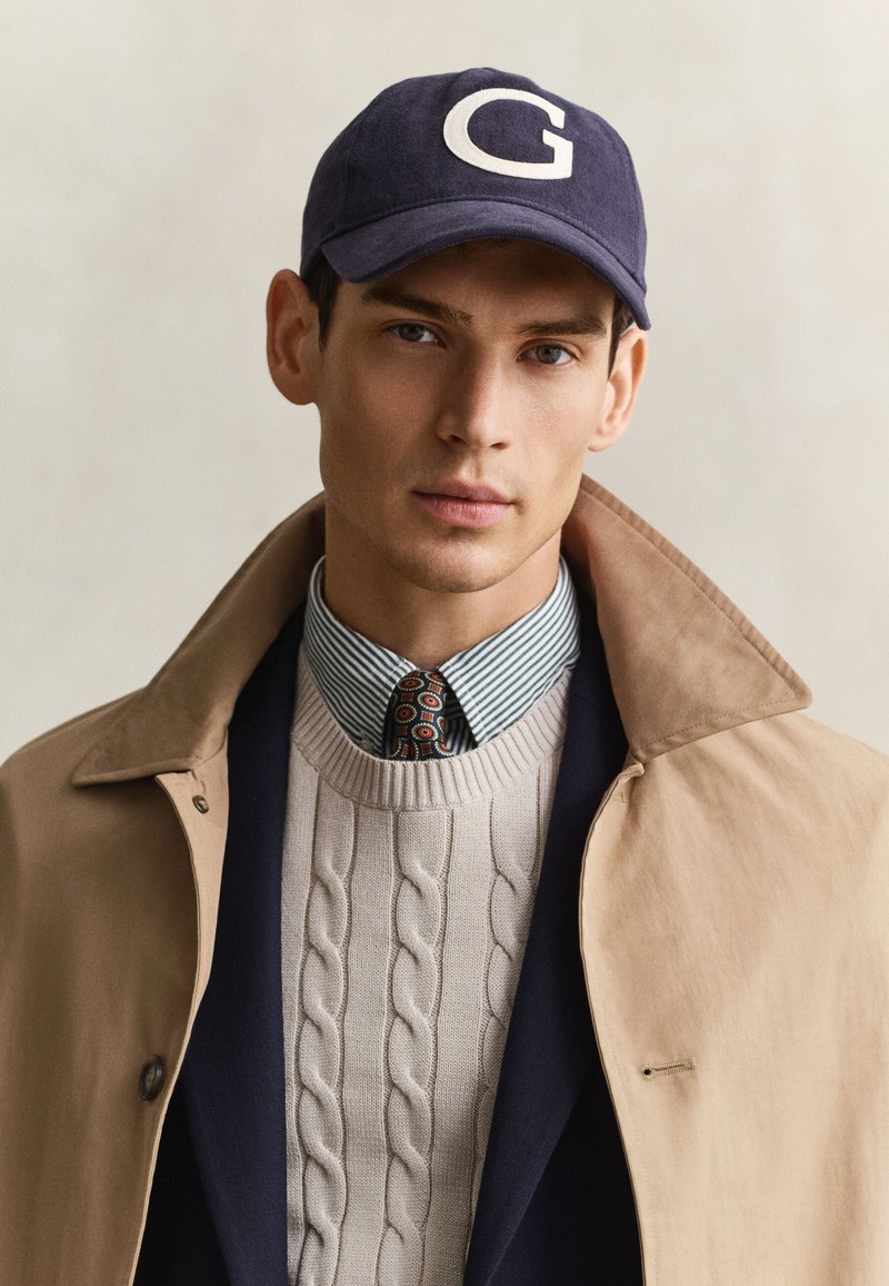 Jeune homme portant un trench beige, une casquette de baseball bleu marine avec un G, un pull léger à torsades, une chemise à rayures et une cravate à motifs.