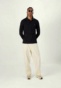 Svart langermet polo-skjorte med en subtil logo, kombinert med kremfargede vide bukser og beige sneakers; satt mot en lys bakgrunn.