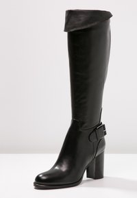 Botte haute en cuir noir jusqu'au genou avec revers plié, talon bloc, bout arrondi et sangle décorative à boucle près de la cheville.