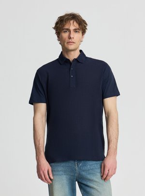 Giovane uomo con capelli castani ondulati che indossa una polo a maniche corte blu scuro e jeans azzurri, in piedi contro uno sfondo semplice.