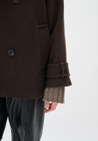 Abrigo de lana marrón con botones de doble botonadura, solapas grandes y puños de punto beige texturizado. Lleva puesto sobre pantalones de cuero negros.