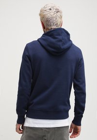 Sudadera con capucha azul marino con cordón ajustable, puños y cinturilla acanalados. Tela suave con un ajuste relajado, combinada con una camisa blanca.