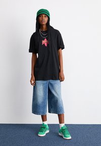 Svart oversized t-shirt med röd grafik, ljusblå vida denimshorts och gröna sneakers med texturerade detaljer. Grönrandig stickad beanie.