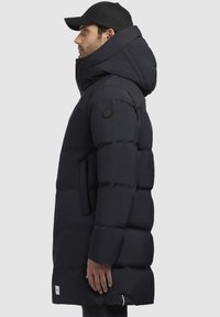 Giacca puffer blu navy con colletto alto e cappuccio grande, dotata di una toppa con logo sulla manica e di una tasca laterale con zip. Tessuto liscio.