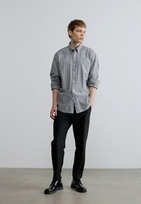 Szürke és fehér csíkos, gombos hosszú ujjú ing fekete nadrággal és fekete cipővel, semleges háttér előtt állva.