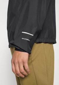 Gros plan d'une personne portant une manche de veste Windwall noire et un pantalon vert olive, main détendue le long du corps.