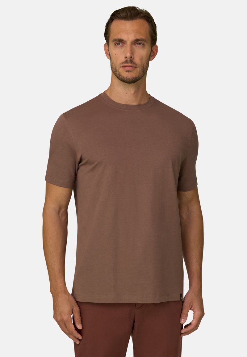 Homme aux cheveux courts et bruns avec une barbe, portant un t-shirt marron uni à manches courtes et un pantalon marron, se tenant devant un fond uni.
