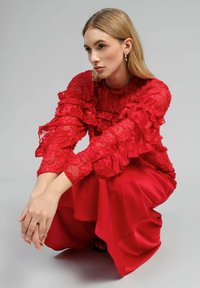 Blouse rouge à volants avec une texture transparente, associée à une jupe rouge fluide. La tenue présente des volants superposés et un éclat lisse.