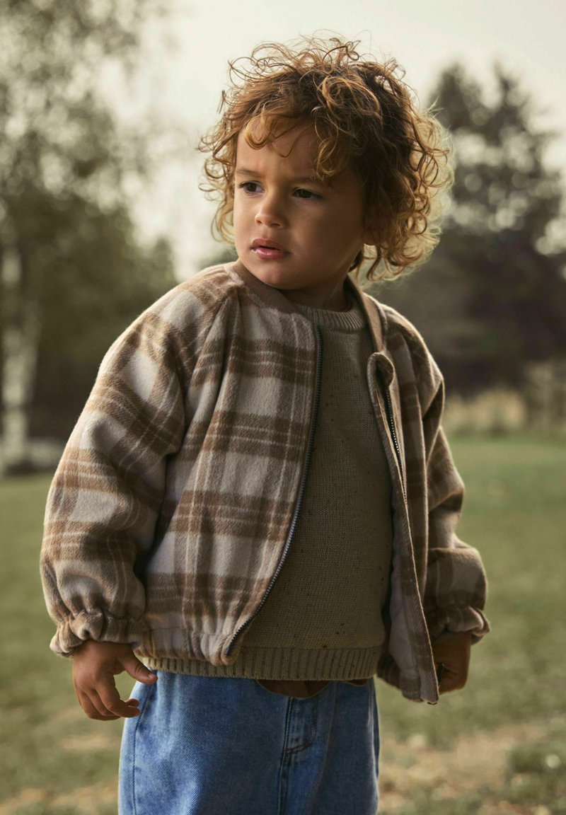Jeune enfant aux cheveux bouclés portant une veste à carreaux beige, un pull et un jean bleu, debout en extérieur dans une zone herbeuse avec des arbres en arrière-plan.
