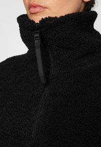 Pullover in pile nero con colletto alto, dotato di zip a metà, superficie a trama e dettaglio della zip in metallo liscio.