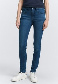 Jeans ajustados de color azul, hechos de denim con una textura suave, que presentan un cierre frontal con botón y bolsillos laterales, combinados con zapatillas blancas.